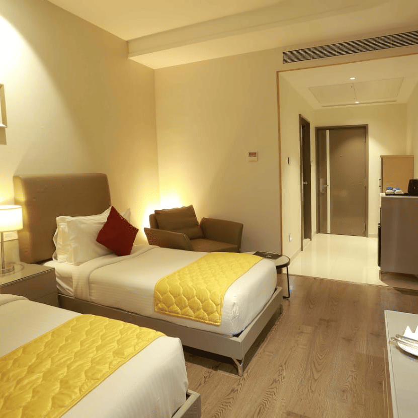 Premium Twin Room at Mayberry Tiruppur featuring a TV stand, wall-mounted TV, and a view of the outside door.